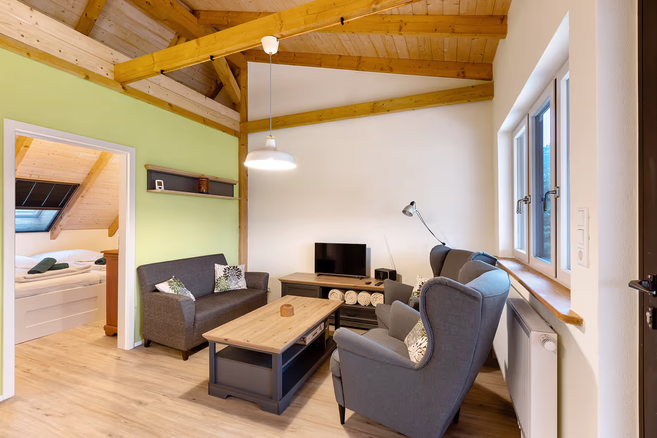 Ein gemütliches Wohnzimmer mit einer hellgrünen Wand und hölzernen Dach. Im Mittelpunk steht eine graue Sofagarnitur mit Tisch